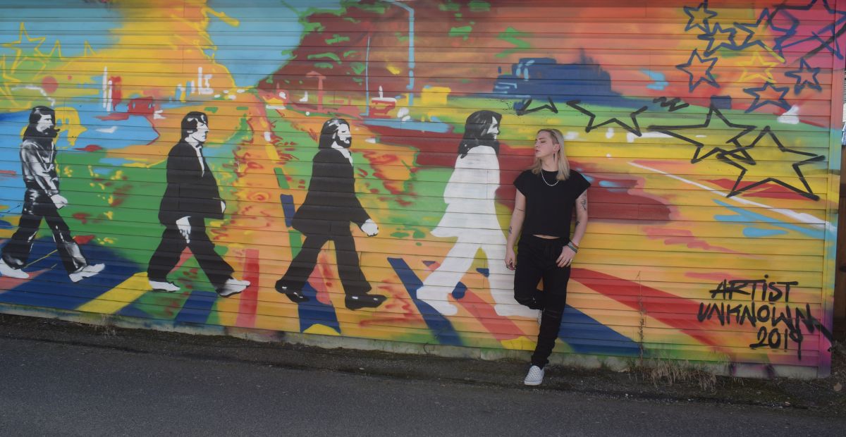 Outdoor mural of the Beatles in black and white in front of a rainbow city scape. Model stands in front of the mural with eyes closed and head turned to the left. Model is wearing black t-shirt, black pants, and silver necklace.