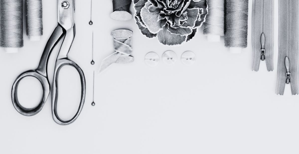 Thread, scissors, pins, flower, buttons, more thread, and zippers lined up in a row. Picture is in black and white.