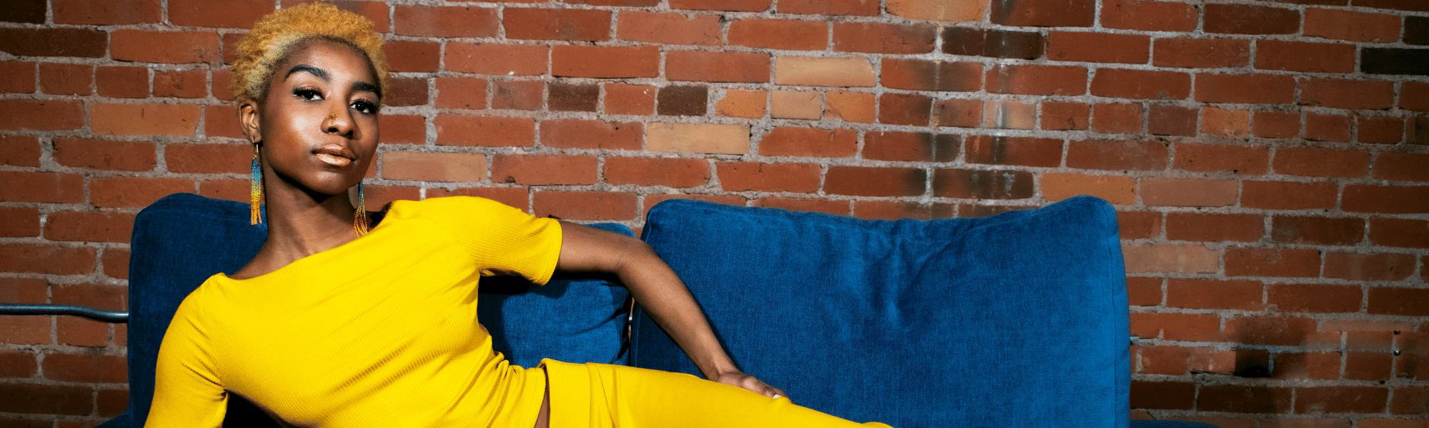 Black model with beaded earrings lounges on a blue velvet couch. The model is wearing a yellow crop top and skirt, and multicolored beaded earrings. They are looking into the camera with a calm and confident gaze.